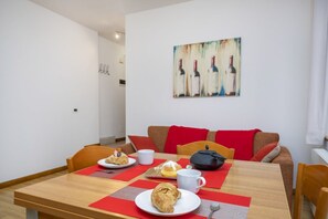 Un comedor con una mesa preparada para dos personas, un cuadro de botellas de vino en la pared y un sofá rojo y marrón al fondo.