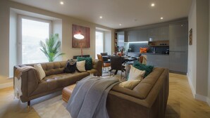 A modern living room with a brown leather sofa, a dining table, and a kitchen area with grey cabinets.