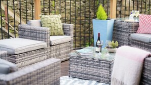A wicker outdoor seating area with cushions, a glass table with a book and a bottle, and a potted plant.