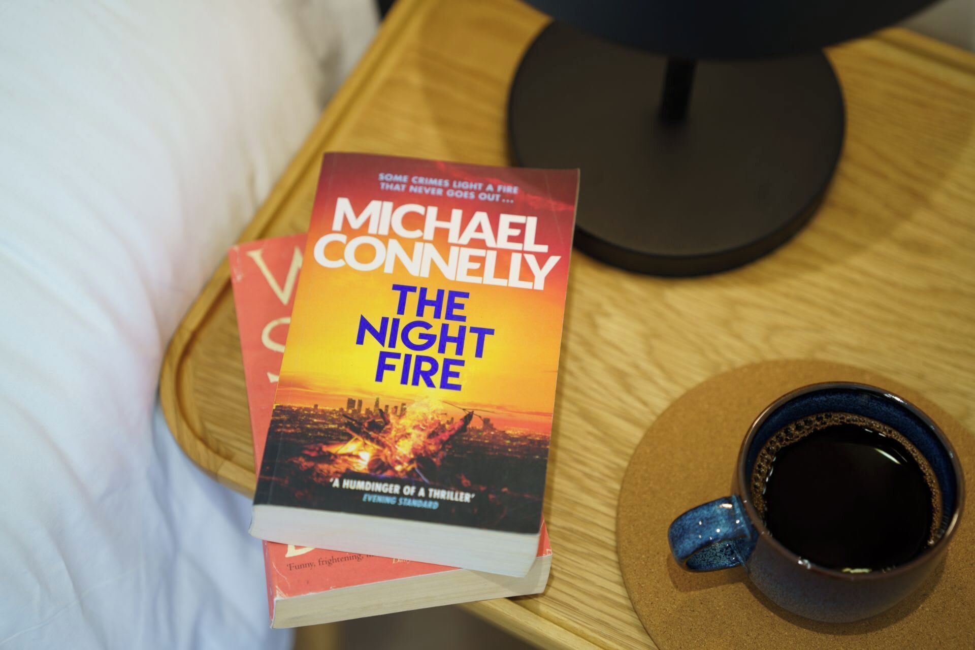 A book titled "The Night Fire" by Michael Connelly on a bedside table next to a cup of coffee.