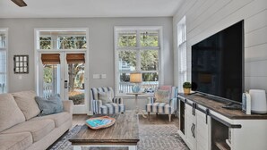 En moderne stue med en sofa, to stribede lænestole, et sofabord og et fjernsyn. Store vinduer med persienner, en loftventilator og et køkkenområde med fjernsyn og skabe.
