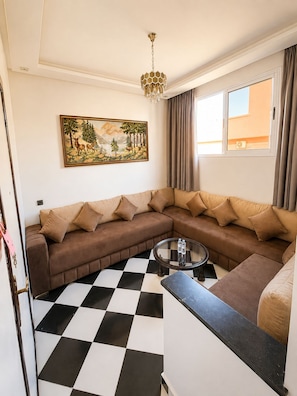 A living room with a brown sectional sofa, a glass coffee table, a checkered floor, a painting of a landscape, and a window with curtains.