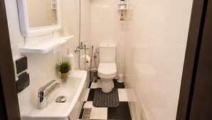 Une petite salle de bain avec un lavabo blanc, un miroir, des toilettes et une douche. Des plantes sont disposées sur les étagères et le sol est carrelé de motifs à carreaux.