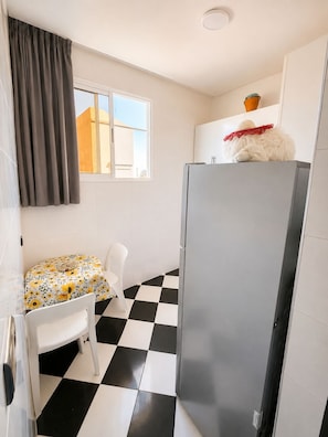 A compact kitchen with a checkered floor, a refrigerator, a small table with a floral tablecloth, and a window with curtains.