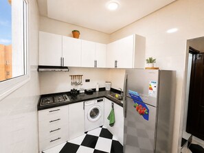 Eine kleine Küche mit weißen Schränken, einer schwarzen Arbeitsplatte, einem Edelstahlkühlschrank, einer Waschmaschine und einem Fenster für natürliches Licht.