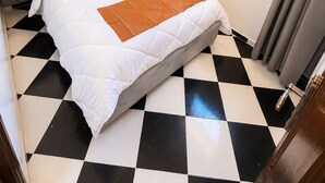 Une chambre d’hôtel avec un sol à carreaux, un lit avec une literie blanche et un plaid marron, une tête de lit grise et une fenêtre avec des rideaux.