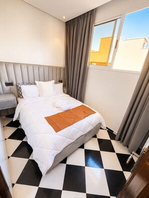 Ein Hotelzimmer mit Schachbrettboden, einem Bett mit weißer Bettwäsche und einer braunen Tagesdecke, einem grauen Kopfteil und einem Fenster mit Vorhängen.
