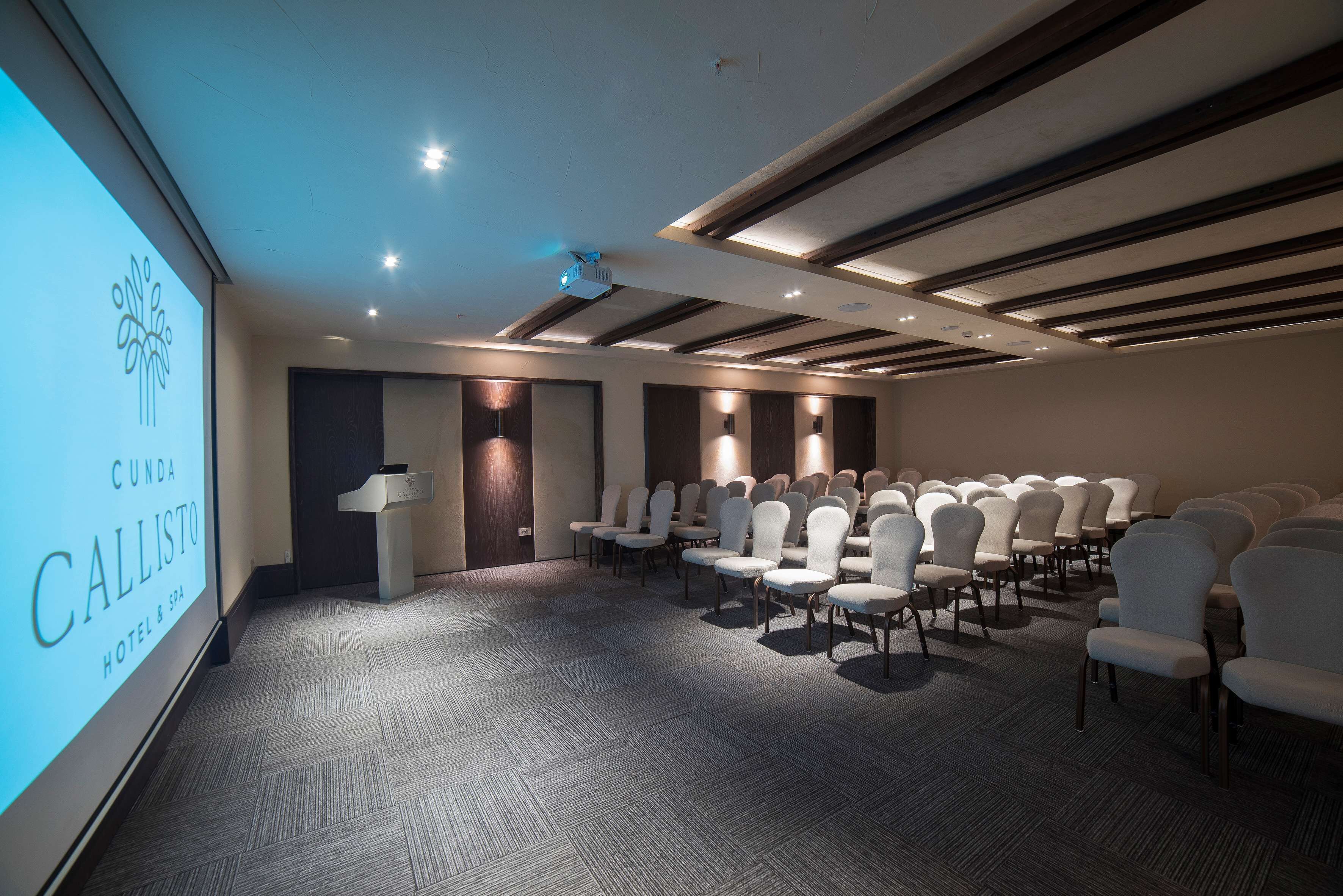 A conference room with rows of white chairs, a podium, and a projection screen displaying "CUNDA CALLISTO HOTEL & SPA".