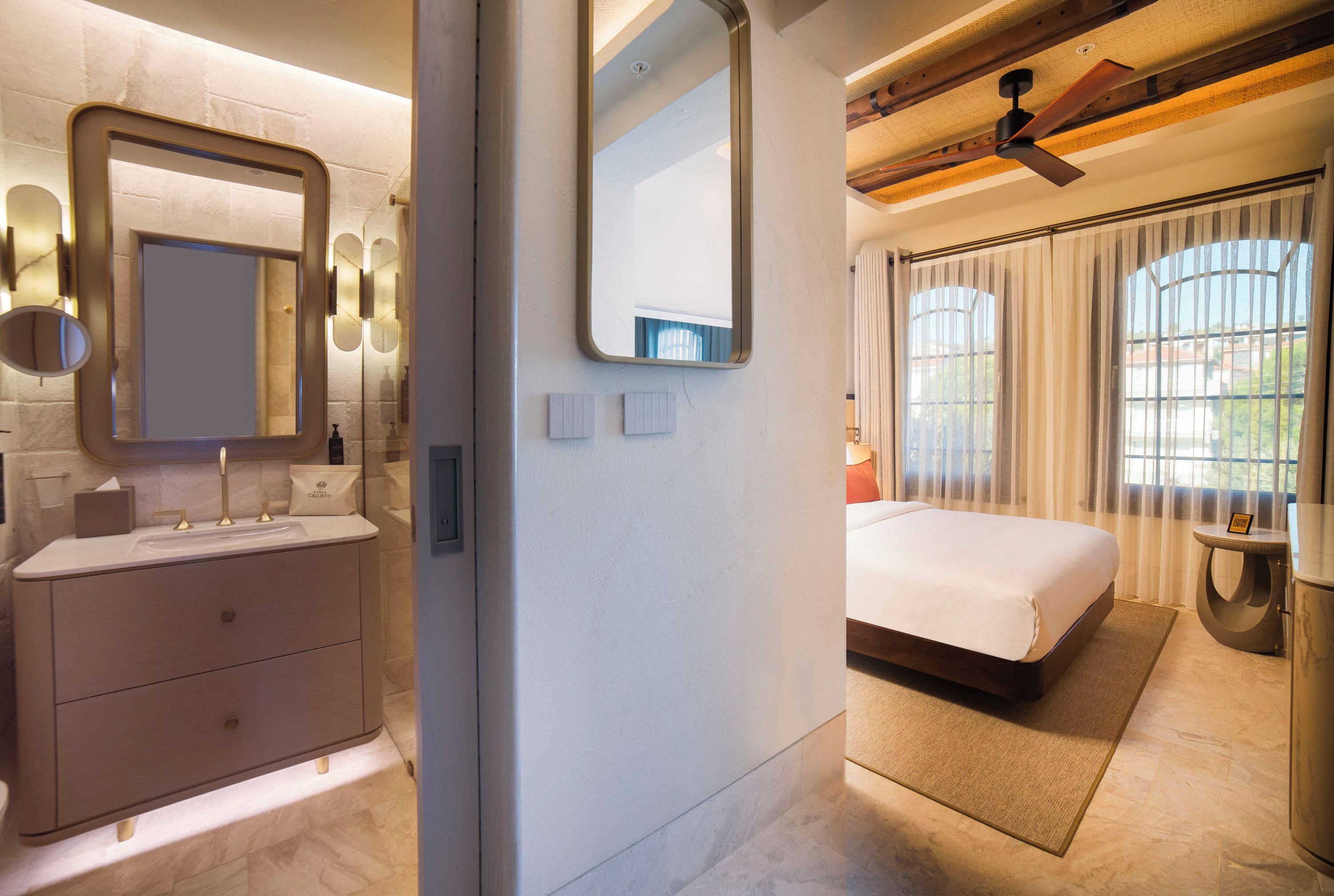 A modern bathroom with a large mirror, a vanity with a sink, and a toilet. The room has a neutral color palette and features a large bed with a view of the outdoors through a window.