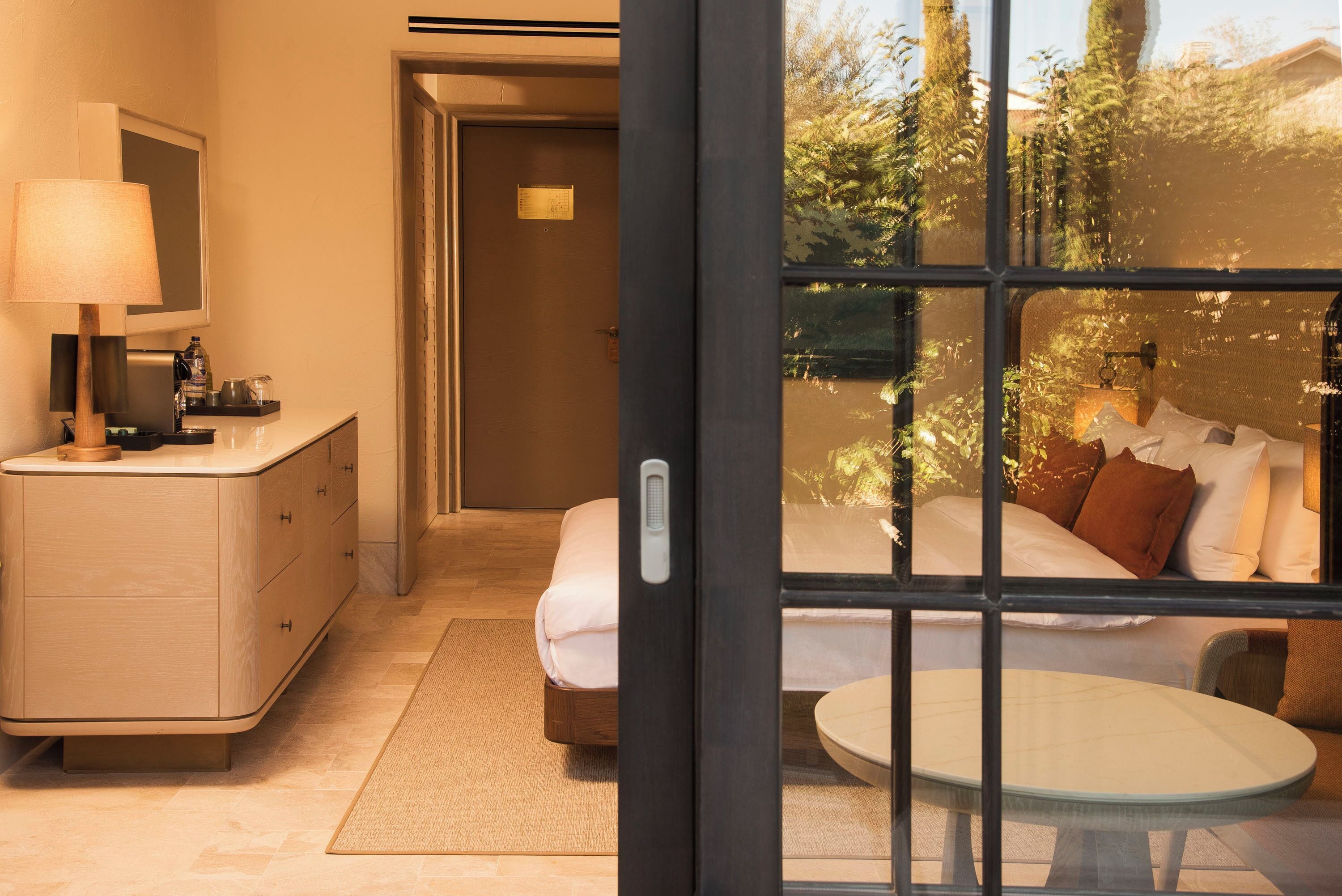 A hotel room with a bed, bedside table, and a window with a view of greenery.