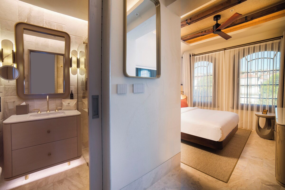 A modern bathroom with a large mirror, a vanity with a sink, and a toilet. The room has a neutral color palette and features a large bed with a view of the outdoors through a window.