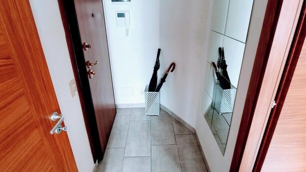Un couloir étroit avec une porte en bois sur la gauche, un mur blanc avec une petite prise électrique et un mur miroir sur la droite. Deux paniers blancs sont posés sur le sol, et chacun contient un parapluie.