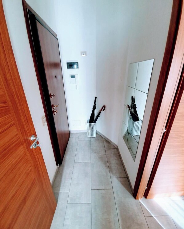 A narrow hallway with a wooden door on the left, a white wall with a small electrical outlet, and a mirrored wall on the right. There are two white baskets on the floor, each holding an umbrella. - Milan shopping street house (Milano)