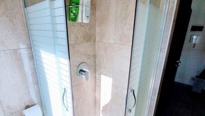 Une douche fermée par des parois en verre, dotée d’un pommeau de douche à main, d’un distributeur de savon en verre et d’un porte-rouleau de papier toilette fixé au mur.