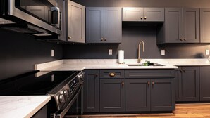 A modern kitchen with dark cabinetry, a stainless steel range hood, and a built-in oven. - The Lilliston - Short Stays on Market (Onancock)
