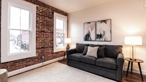 A living room with a brick wall, a gray sofa, two windows, and abstract art on the wall. - The Lilliston - Short Stays on Market (Onancock)