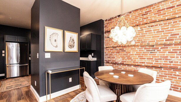 A modern kitchen with a stainless steel refrigerator, a dark wall with framed artwork, and a brick wall with a hanging light fixture. - The Lilliston - Short Stays on Market (Onancock)