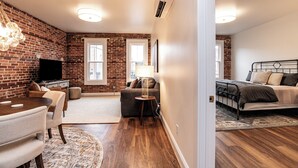 A modern living room with a brick wall, a flat-screen TV, a sofa, a dining table, and a small side table. - The Lilliston - Short Stays on Market (Onancock)