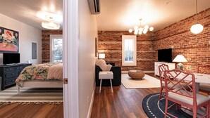 A modern bedroom with a brick wall, wooden flooring, a bed with floral bedding, a desk with a lamp, and a dining area with red chairs.