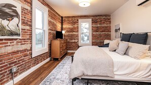 A bedroom with a brick wall, a bed with white bedding, a wooden dresser, and a window.
