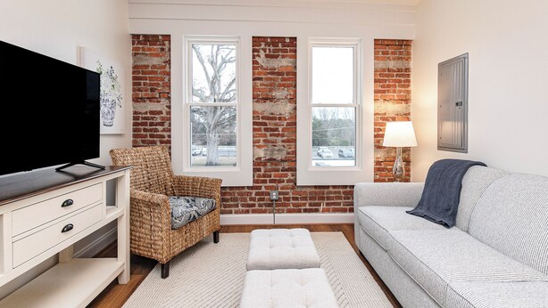 A living room with a brick wall, two windows, a television, a wicker chair, a sofa, and a lamp.