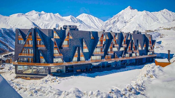 A modern building with a unique geometric design, featuring multiple levels and large windows, set against a snowy mountainous backdrop. - Comfy Studio in Gudauri Marshall - By Wehost (Gudauri)