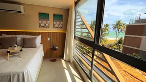 Une chambre d’hôtel avec un lit, une petite table sur laquelle se trouvent une tasse et une soucoupe, une plante décorative et une grande fenêtre donnant sur une plage et des palmiers.