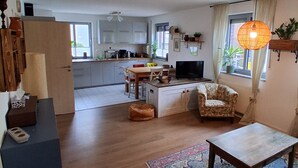 Een ruime woonkamer met een bank, een salontafel en een kamerkleed. In de achtergrond is een keuken met witte kasten en een eethoek met een tafel en stoelen zichtbaar.