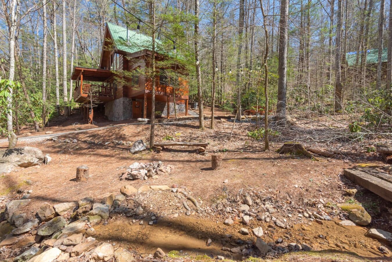 A rustic cabin with a green roof, surrounded by a forested area with a dirt path leading to it.