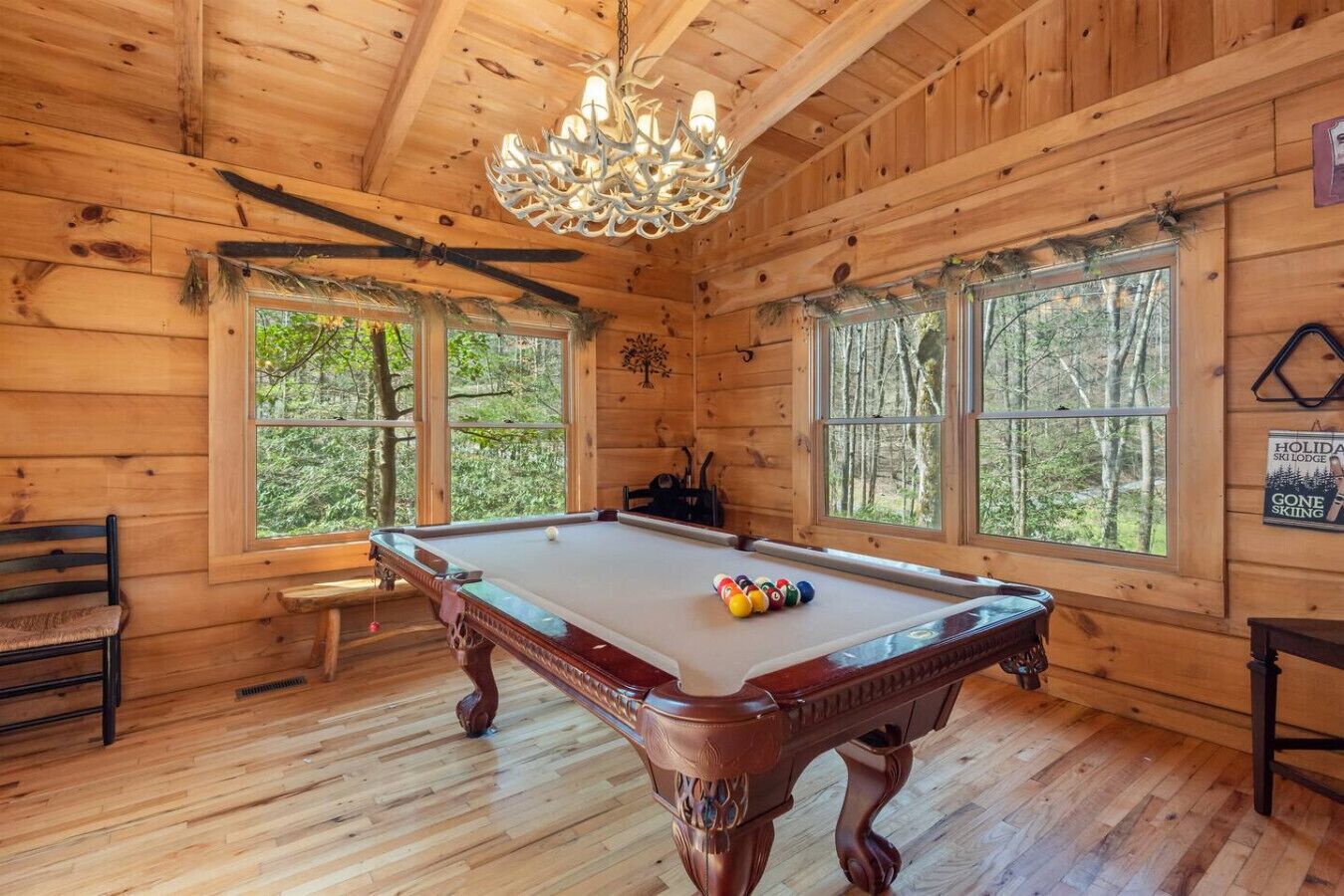 A wooden cabin with a pool table, benches, and large windows offering a forest view.