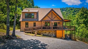 A two-story log cabin with a stone foundation, a wooden deck, and a view of a forested hillside. - Luxe 4BR Pool Cabin*Hot Tub*Firepit*Games*Theater! (Sevierville)