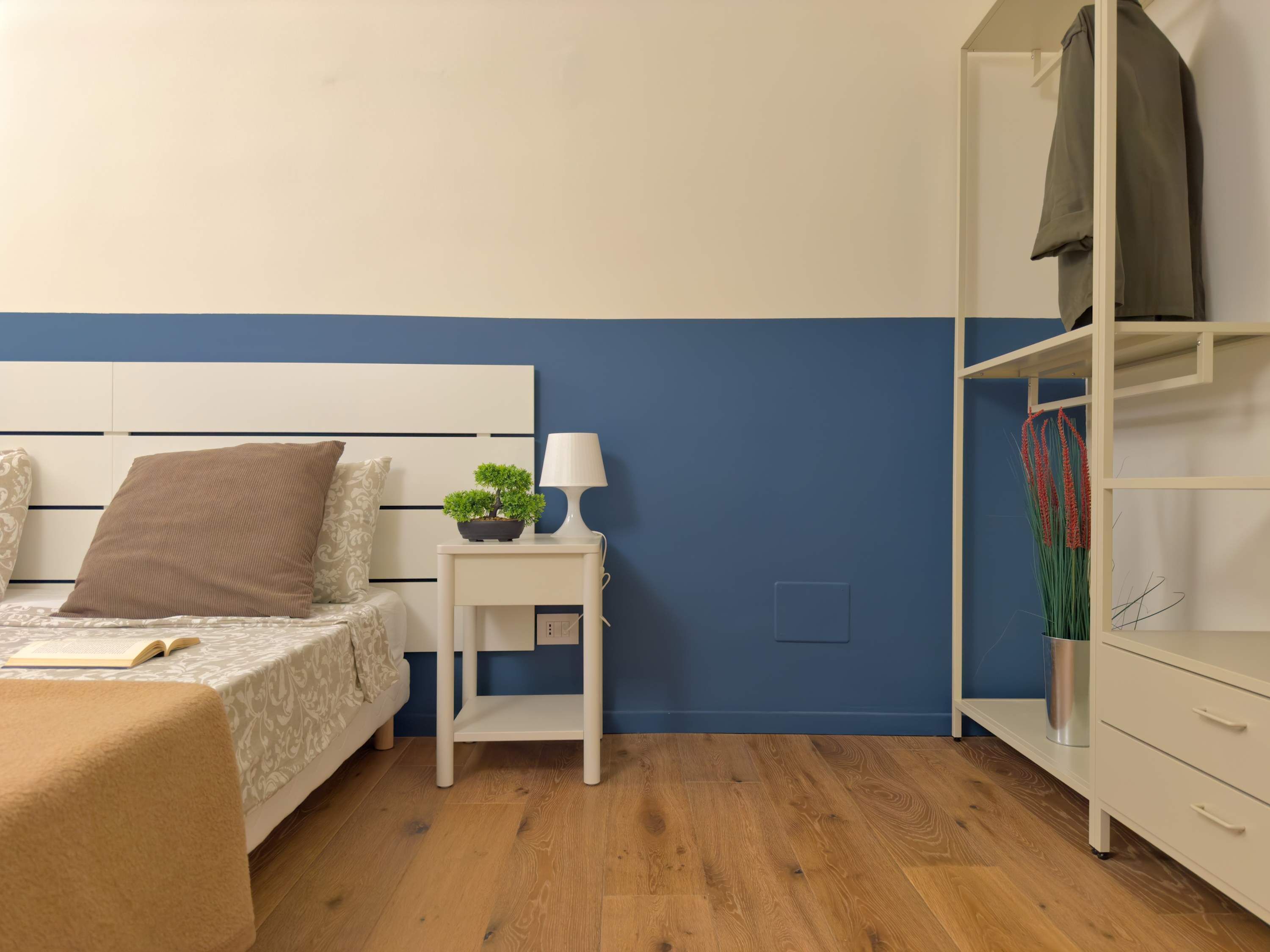 A bedroom with a bed, bedside table with a lamp, a small potted plant, and a wardrobe.