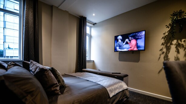 A modern hotel room with a large bed, a flat-screen TV mounted on the wall, a window with curtains, and a plant on the right side. - Ladywell House (Birmingham)