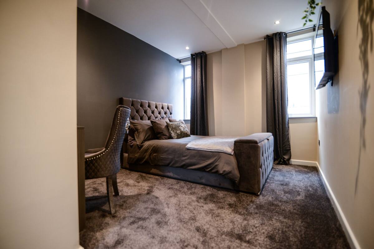 A bedroom with a tufted headboard bed, a chair, a large window with curtains, and a television on the wall.