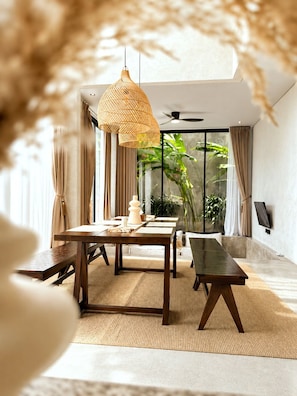 A modern dining area with a large table, benches, a hanging light fixture, and a view of greenery through large windows. - Industrial Style 2 Bedroom Villa with Private Pool in Pererenan (Badung Regency)
