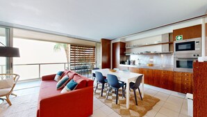 A modern kitchen with stainless steel appliances, a dining area with a red sofa and blue chairs, and a view of palm trees through large windows. - Herdade dos Salgados, T2 12C_2A, Vila das Lagoas (Albufeira)
