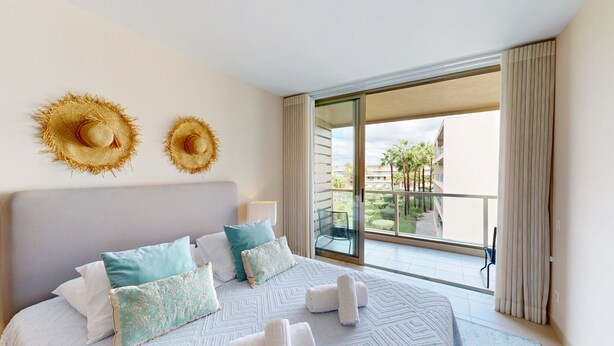 A hotel room with a bed, two wall-mounted hats, a balcony with a view of palm trees and buildings, and a sliding glass door. - Herdade dos Salgados, T2 12C_2A, Vila das Lagoas (Albufeira)