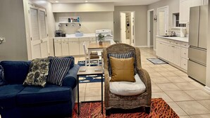 A modern kitchen with white cabinets, a dining area with a wicker chair, and a blue sofa with patterned cushions.