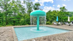 Une piscine dotée d’une fontaine centrale, entourée de chaises longues et de parasols, le tout se fondant dans un décor composé d’arbres et d’un ciel dégagé.