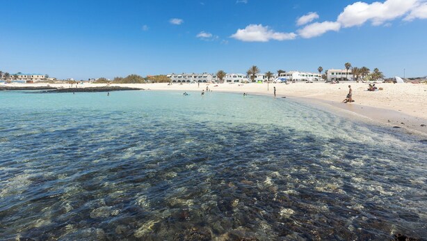 Ein Strand mit klarem Wasser, sandigem Ufer und Gebäuden im Hintergrund.