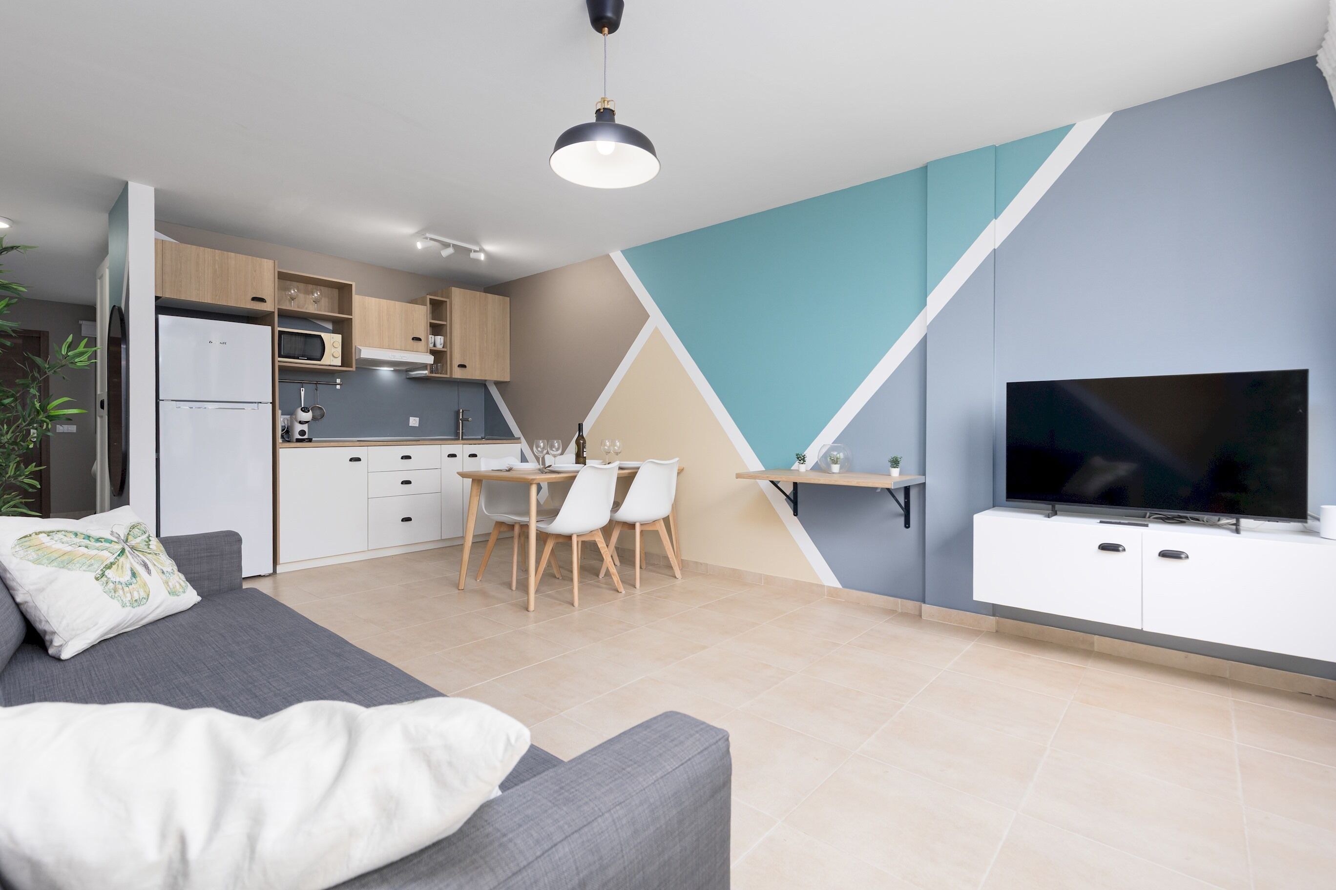 Ein modernes Wohnzimmer mit grauem Sofa, einem Essbereich mit Tisch und Stühlen, einer Küche mit Holzausstattung und einem an einer weißen Kommode befestigten Flachbildfernseher.