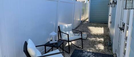 Un espace patio comprenant deux chaises blanches avec coussins, une petite table et un chemin menant à un bâtiment doté d’une porte bleue.