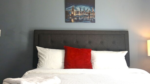 Una cama bien hecha con ropa de cama blanca y un cojín rojo, una obra de arte urbana en un marco en la pared y un cabecero oscuro.