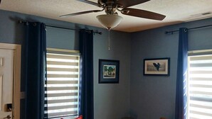 Una sala de estar con un ventilador de techo, dos ventanas con persianas, un sillón blanco con un cojín rojo, un sofá naranja con cojines rojos y amarillos, y una mesa de centro con un control remoto.
