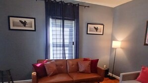 Una sala de estar con un sofá marrón, una mesita de centro redonda y un sillón blanco. Hay dos cuadros enmarcados en la pared, una ventana con cortinas y una lámpara de pie.