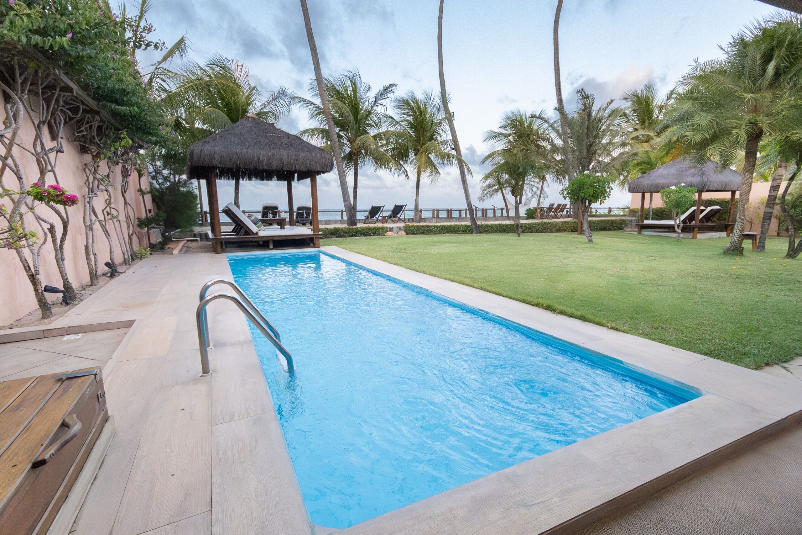A swimming pool with lounge chairs and a thatched-roof structure, surrounded by palm trees and a well-maintained lawn.