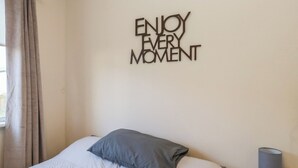 A bedroom with a bed, a wall-mounted sign saying "ENJOY EVERY MOMENT," a lamp, and a window with curtains. - Beechtree Lodge 54 (Denny)