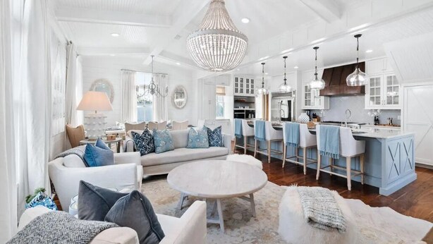 Ein geräumiges Wohnzimmer mit einer großen Couch, einem runden Couchtisch und einem Essbereich mit blauen Stühlen sowie einer Küche im Hintergrund.