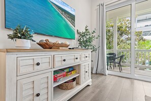 A room with a large window, a white cabinet with drawers, a television displaying a beach scene, and a view of a balcony with outdoor furniture. - ⛱️Beach Access | Pet Friendly | Azure on 30A 1-205- A Wave From it All⛱️ (Santa Rosa Beach)