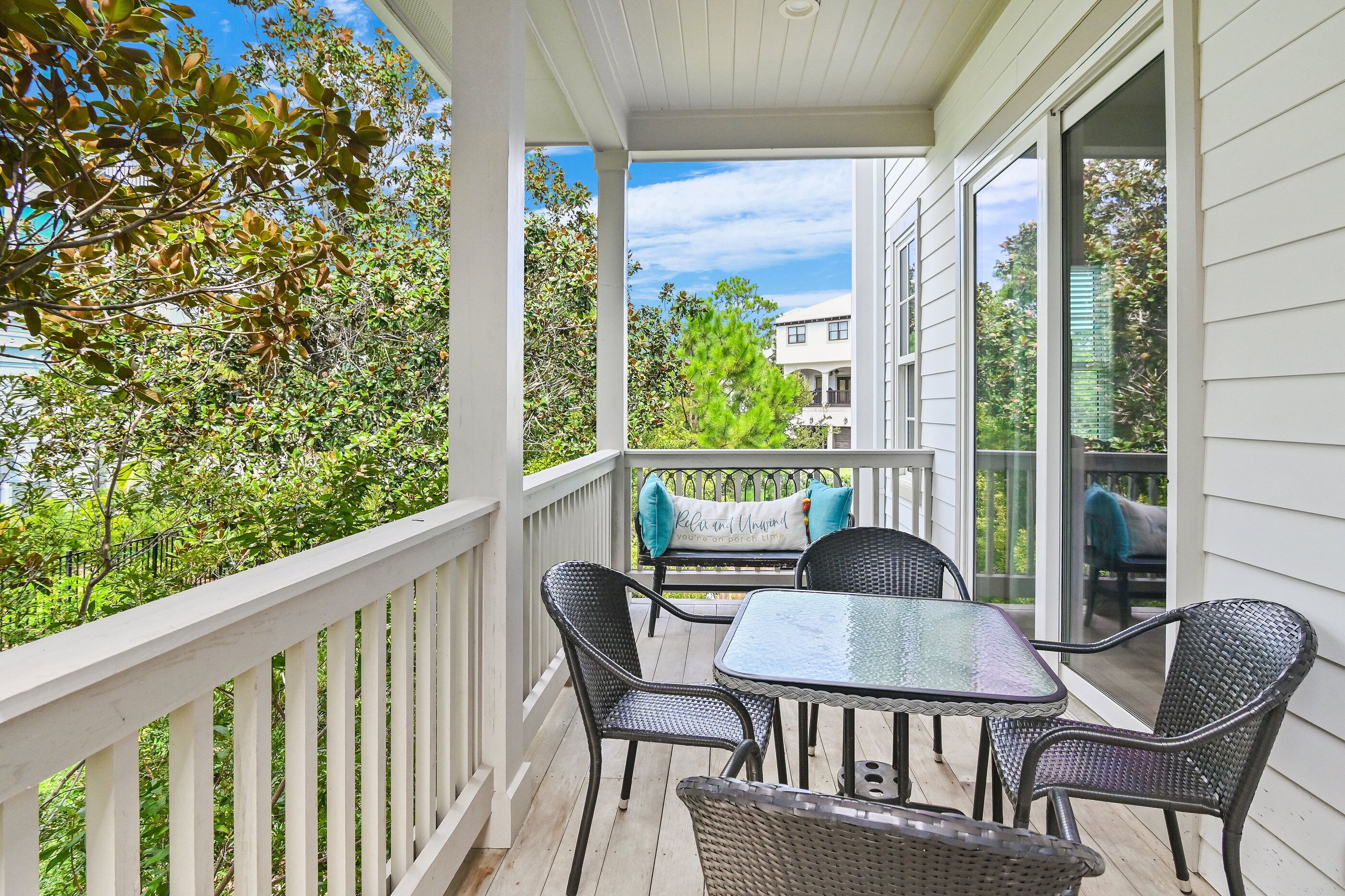 A covered outdoor patio with a table and chairs, a white railing, and a view of a building and trees.